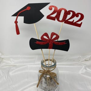 Puede incluir: Centro de mesa de graduaci&oacute;n con un gorro de graduaci&oacute;n negro, un diploma rojo con un lazo rojo y un 2022 rojo con purpurina. Las decoraciones se colocan en un frasco de vidrio transparente con una cinta de arpillera.