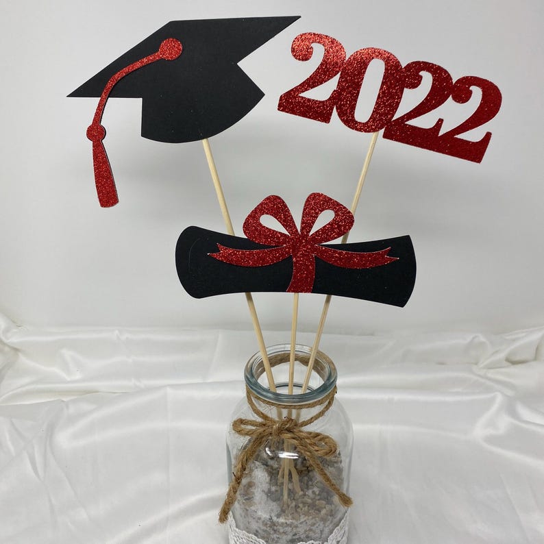 Puede incluir: Centro de mesa de graduaci&oacute;n con un gorro de graduaci&oacute;n negro, un diploma rojo con un lazo rojo y un 2022 rojo con purpurina. Las decoraciones se colocan en un frasco de vidrio transparente con una cinta de arpillera.