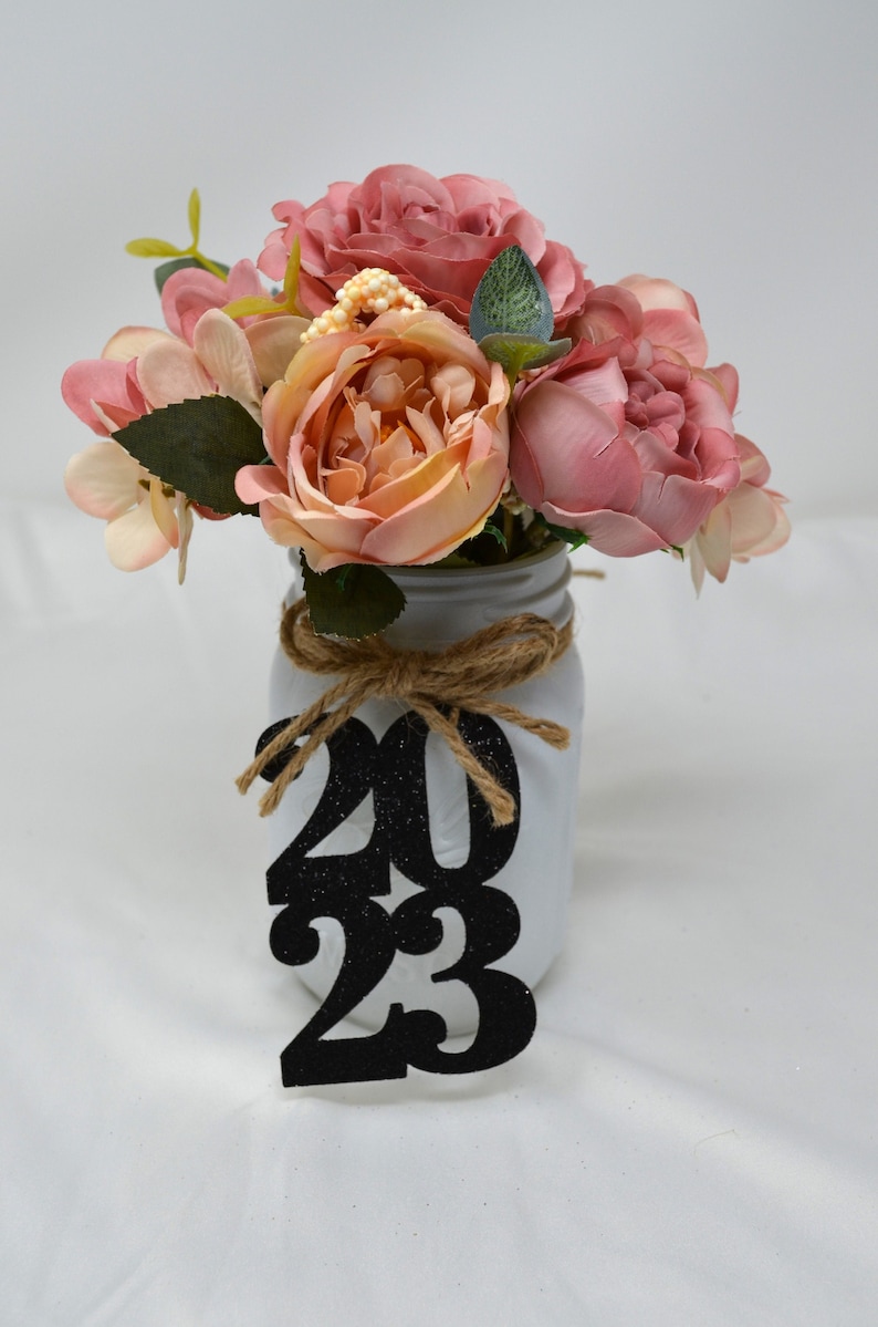 May include: A white mason jar with a burlap bow and a black glitter 2023 sign. The jar is filled with a bouquet of pink and peach artificial flowers.