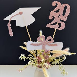 May include: A graduation centerpiece with a white graduation cap, a white diploma with a pink ribbon, and a rose gold glitter 2023 on wooden sticks. The centerpiece is in a small white vase with dried flowers.