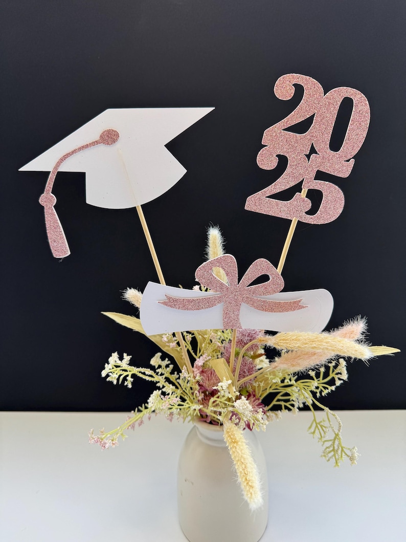 May include: A graduation centerpiece with a white graduation cap, a white diploma with a pink ribbon, and a rose gold glitter 2023 on wooden sticks. The centerpiece is in a small white vase with dried flowers.