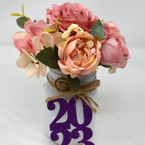 May include: A white mason jar with a burlap bow and a purple glitter 2023 sign. The jar is filled with a bouquet of pink and peach artificial flowers.