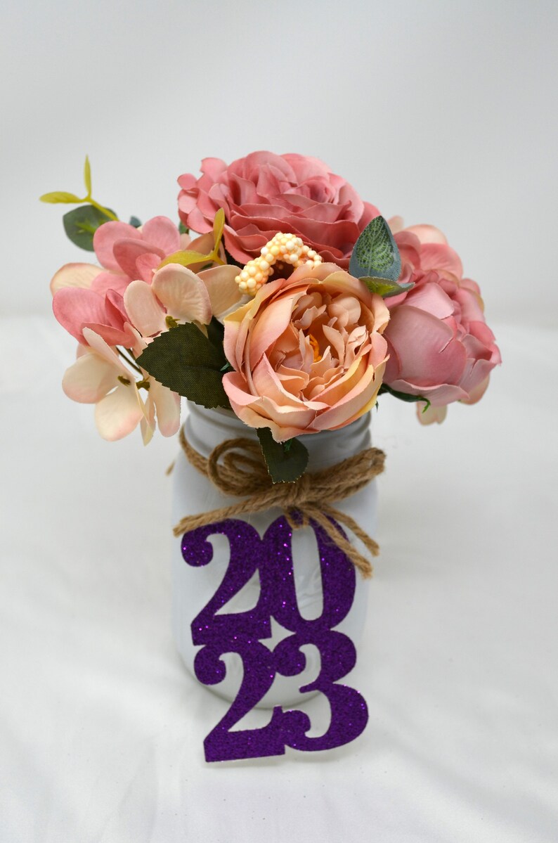 May include: A white mason jar with a burlap bow and a purple glitter 2023 sign. The jar is filled with a bouquet of pink and peach artificial flowers.