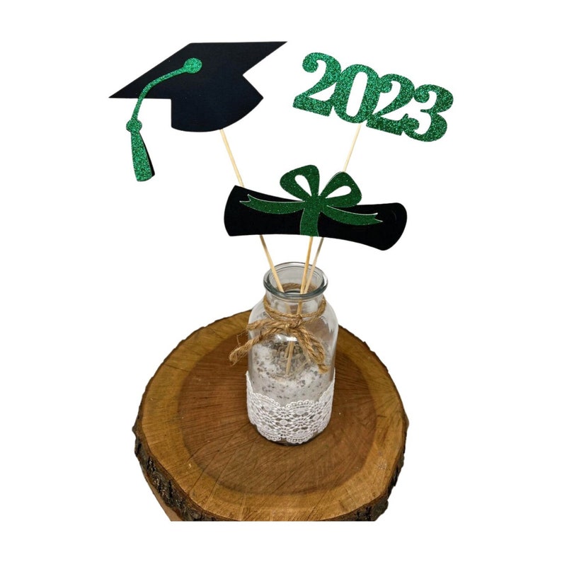 Puede incluir: Centro de mesa de graduaci&oacute;n con un gorro de graduaci&oacute;n negro, un diploma verde y un 2023 verde con purpurina en palos de madera en un jarr&oacute;n de vidrio transparente con un borde de encaje y una cinta de arpillera en una rodaja de madera.