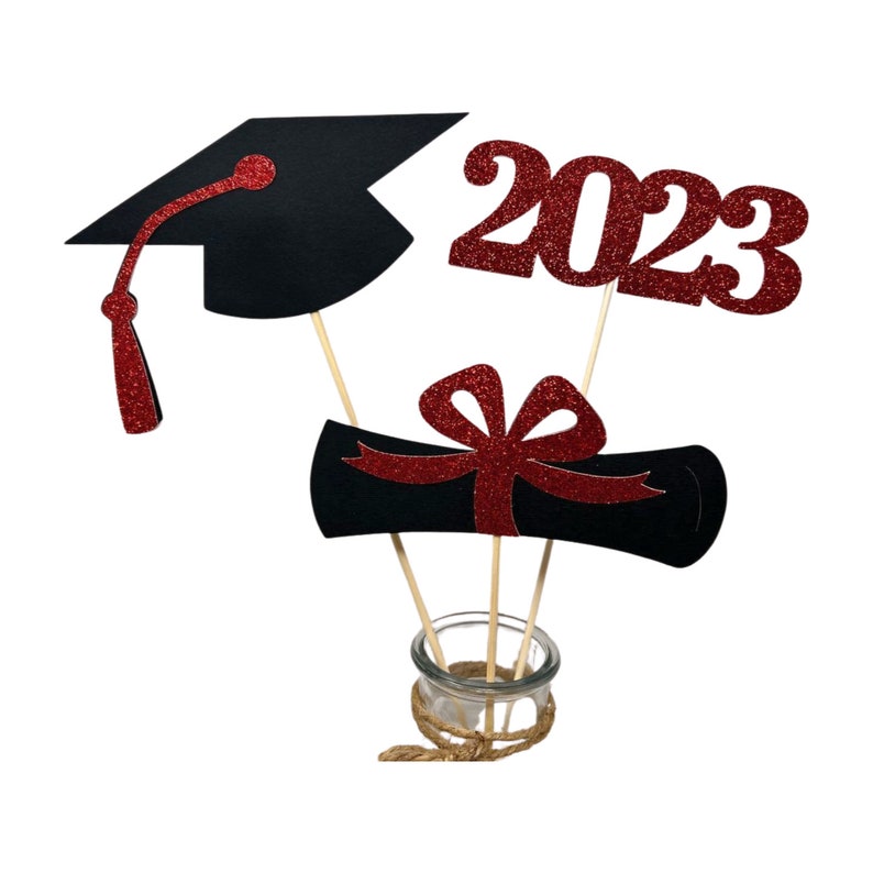 Puede incluir: Centro de mesa para una fiesta de graduaci&oacute;n con un gorro de graduaci&oacute;n negro, un diploma rojo con un lazo y un 2023 de purpurina roja.
