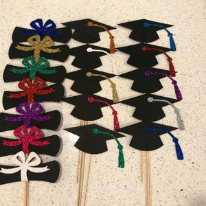 May include: A set of 10 graduation cap and diploma cake toppers. The toppers are made of black cardstock with glitter accents. The caps have different colored tassels and the diplomas have different colored bows.