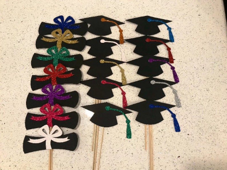 May include: A set of 10 graduation cap and diploma cake toppers. The toppers are made of black cardstock with glitter accents. The caps have different colored tassels and the diplomas have different colored bows.