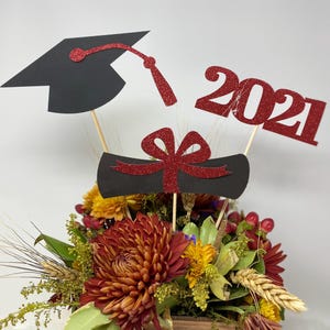 Puede incluir: Centro de mesa de graduaci&oacute;n con un gorro de graduaci&oacute;n negro, un diploma rojo, un lazo rojo y un letrero de 2021 con purpurina roja. El centro de mesa est&aacute; rodeado de una variedad de flores de oto&ntilde;o.