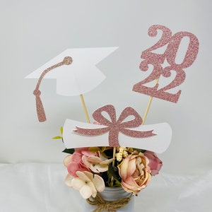 May include: Graduation centerpiece with a white graduation cap, diploma, and rose gold glitter 2022. The centerpiece is in a white mason jar with a burlap bow and pink and white flowers.