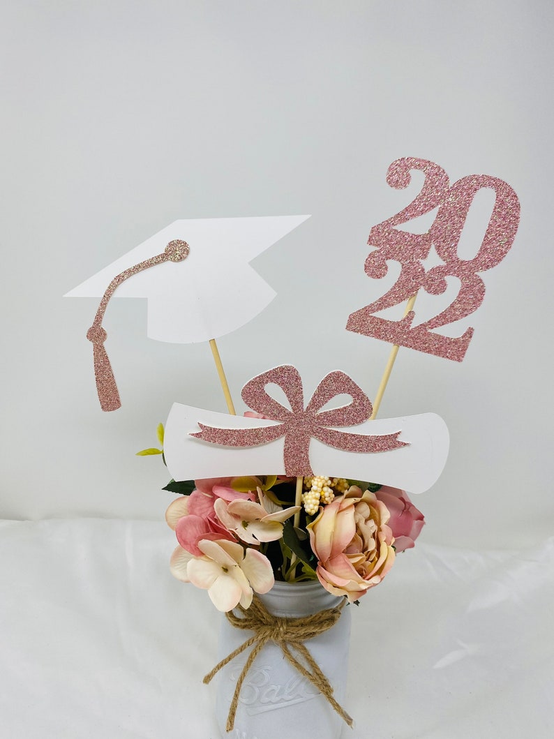 May include: Graduation centerpiece with a white graduation cap, diploma, and rose gold glitter 2022. The centerpiece is in a white mason jar with a burlap bow and pink and white flowers.