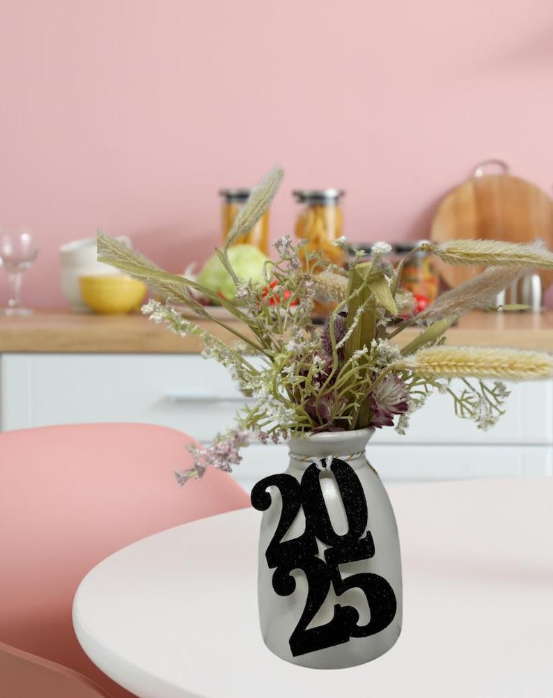 May include: A white ceramic vase with a black glitter 2025 sign attached to it. The vase is filled with dried flowers and grasses.
