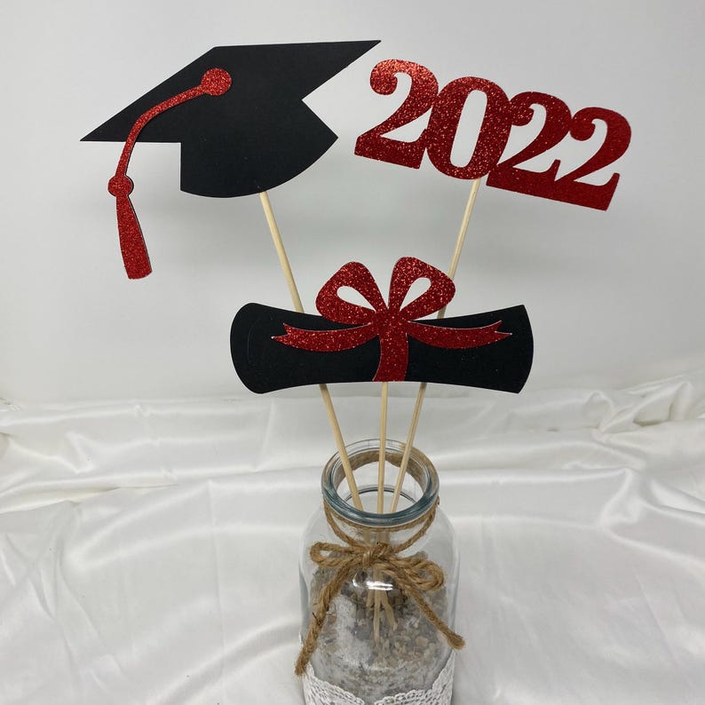 Puede incluir: Centro de mesa de graduaci&oacute;n con un gorro de graduaci&oacute;n negro, un diploma y un letrero de 2022 con purpurina roja. Las decoraciones est&aacute;n en palos de madera en un frasco de vidrio transparente.