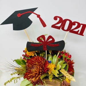 Puede incluir: Un centro de mesa de graduaci&oacute;n con un gorro de graduaci&oacute;n negro, un rollo de diploma rojo y negro y un letrero de brillo rojo 2021. El centro de mesa est&aacute; rodeado de una variedad de flores de oto&ntilde;o en una caja de madera.