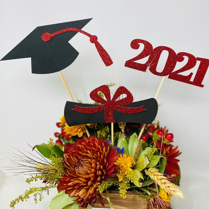 Puede incluir: Un centro de mesa de graduaci&oacute;n con un gorro de graduaci&oacute;n negro, un rollo de diploma rojo y negro y un letrero de brillo rojo 2021. El centro de mesa est&aacute; rodeado de una variedad de flores de oto&ntilde;o en una caja de madera.