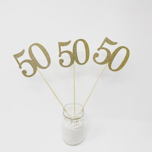 May include: Three gold glitter '50' numbers on sticks in a clear glass jar filled with white decorative rocks.