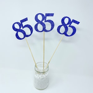 May include: Three blue glitter number 85s on wooden sticks in a clear glass jar with white rocks.