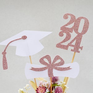 May include: Graduation centerpiece with a white graduation cap, a white diploma scroll with a pink bow, and a pink glitter 2024.
