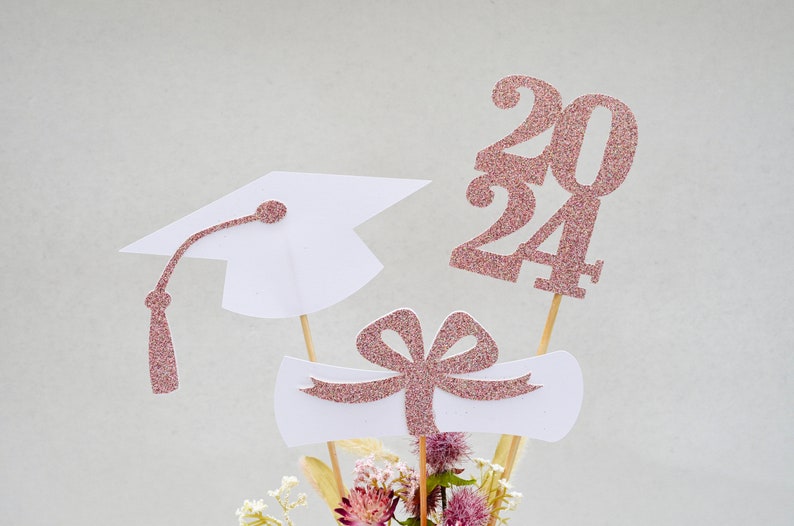 May include: Graduation centerpiece with a white graduation cap, a white diploma scroll with a pink bow, and a pink glitter 2024.