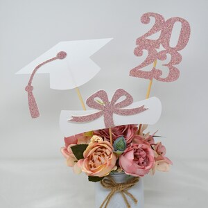 May include: A graduation centerpiece with a white mason jar, pink flowers, and pink glitter graduation cap, diploma, and the year 2023.