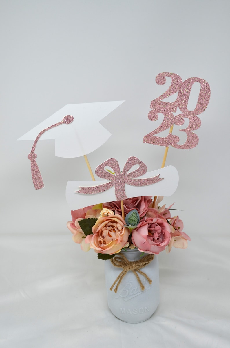 May include: A graduation centerpiece with a white mason jar, pink flowers, and pink glitter graduation cap, diploma, and the year 2023.