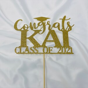 May include: Gold glitter cake topper with the text "Congrats Kai Class of 2021" and a graduation cap.