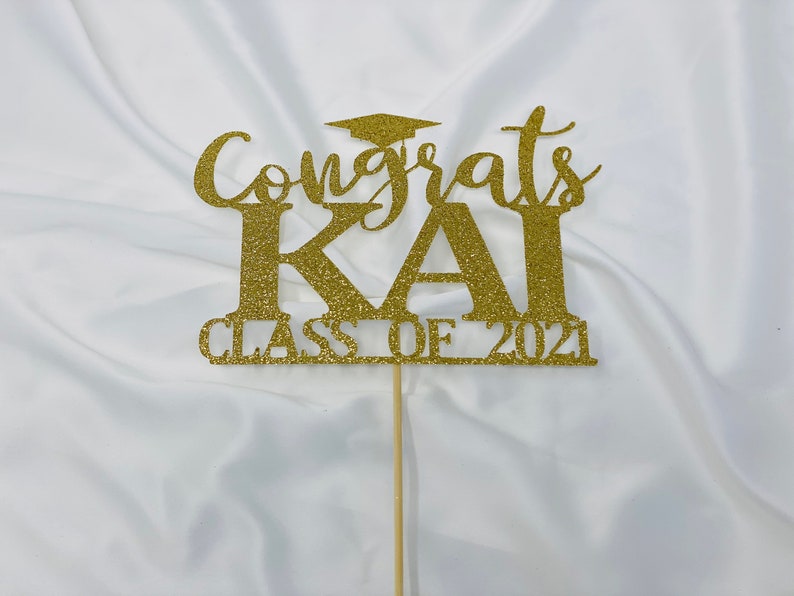 May include: Gold glitter cake topper with the text "Congrats Kai Class of 2021" and a graduation cap.