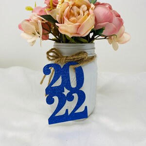 May include: A white mason jar with a burlap ribbon and a blue glitter 2022 sign. The jar is filled with a bouquet of pink and white flowers.