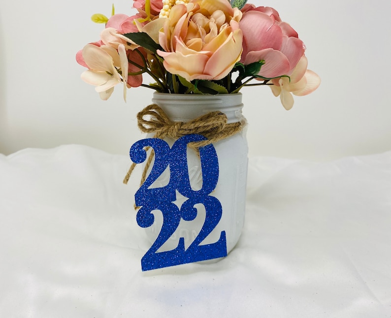 May include: A white mason jar with a burlap ribbon and a blue glitter 2022 sign. The jar is filled with a bouquet of pink and white flowers.
