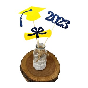 Puede incluir: Centro de mesa de graduaci&oacute;n con un gorro de graduaci&oacute;n amarillo, un diploma y 2023 en purpurina azul. Las decoraciones se colocan en un jarr&oacute;n de vidrio transparente con un borde de encaje y un lazo de arpillera. El jarr&oacute;n est&aacute; colocado sobre una rodaja de madera.