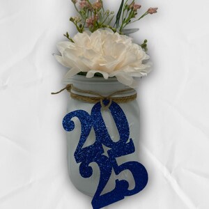 May include: A white mason jar with a blue glitter 2025 tag tied around the top. The jar is filled with a white flower and pink flowers.
