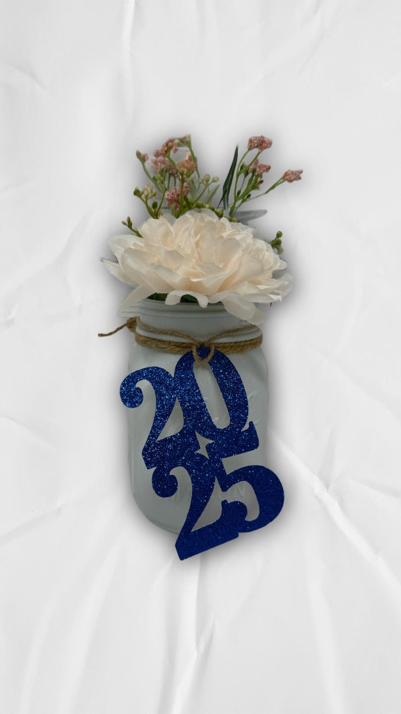 May include: A white mason jar with a blue glitter 2025 tag tied around the top. The jar is filled with a white flower and pink flowers.