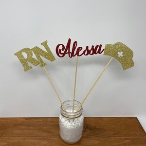 May include: Gold glitter "RN" and "Alessa" with a red glitter nurse's hat on wooden sticks in a clear glass jar with white rocks.