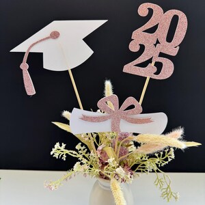 May include: Graduation centerpiece with a rose gold glitter graduation cap, diploma, and the year 2023. The centerpiece is in a small vase with dried flowers.