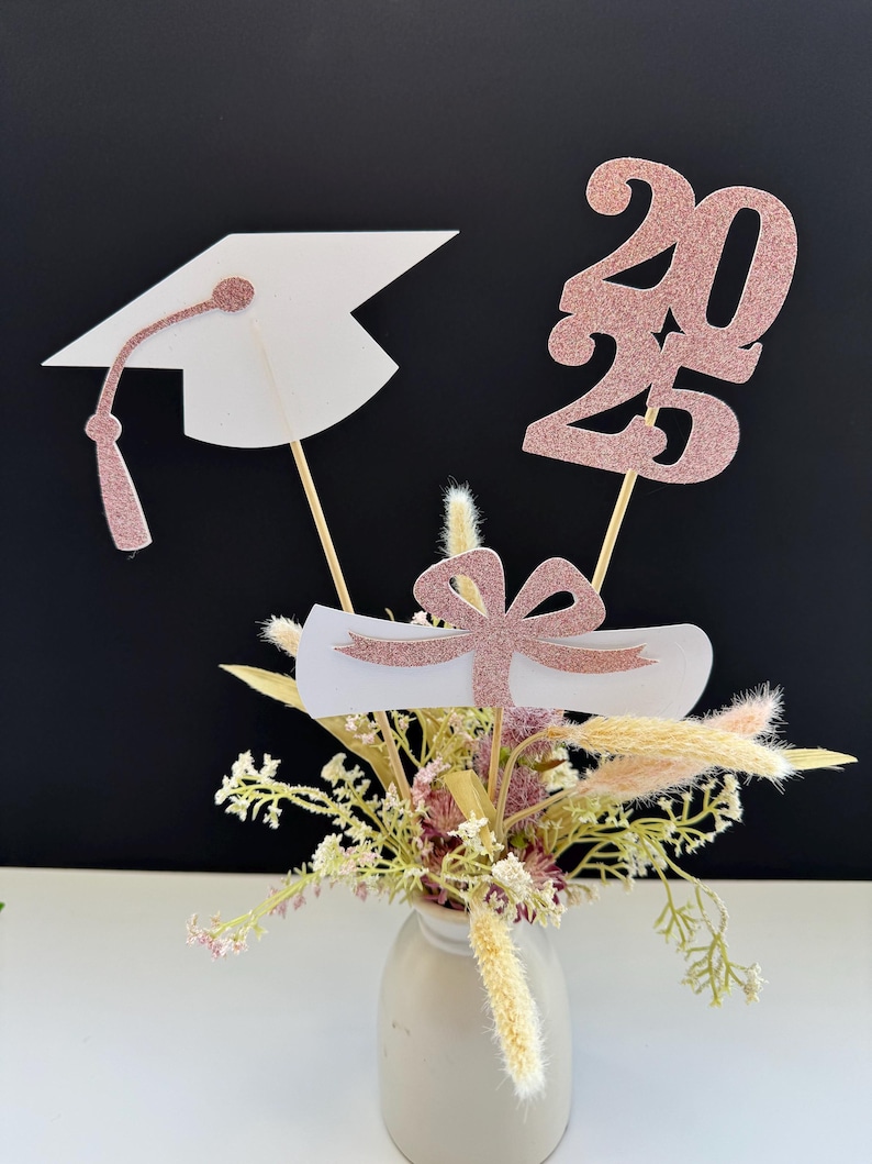 May include: Graduation centerpiece with a rose gold glitter graduation cap, diploma, and the year 2023. The centerpiece is in a small vase with dried flowers.