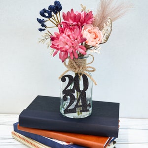May include: A glass vase with a burlap ribbon and a black glitter 2024 sign, filled with pink and peach flowers, blue berries, and dried pampas grass. The vase is sitting on a stack of three books.