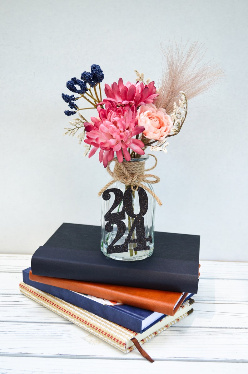 May include: A glass vase with a burlap ribbon and a black glitter 2024 sign, filled with pink and peach flowers, blue berries, and dried pampas grass. The vase is sitting on a stack of three books.