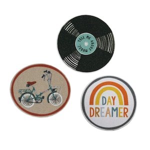 Op de afbeelding: Drie ronde stoffen patches met verschillende ontwerpen. De eerste patch is zwart met een wit platenontwerp en de tekst "Tell Me About Music". De tweede patch is beige met een rode rand en een groene fiets. De derde patch is wit met een grijze rand en een regenboogontwerp met de tekst "Day Dreamer".