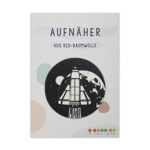 Könnte beinhalten: Schwarz-weißes Bügelbild mit einer Rakete, die an einem Planeten vorbeifliegt. Der Patch trägt die Aufschrift "AUFNÄHER AUS BIO-BAUMWOLLE" und "TELL ME organic goods".