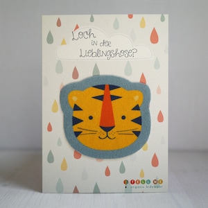 Könnte beinhalten: Eine weiße Karte mit einer blauen und gelben Tigergesichts-Illustration. Die Karte hat eine Wolke mit dem Text "Loch in der Lieblingshose?" und ein buntes Regentropfenmuster. Am unteren Rand der Karte steht der Text "Tell Me organic kidswear."