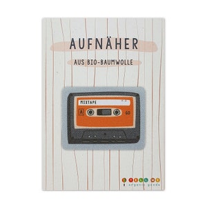 May include: A blue and orange fabric iron-on patch of a cassette tape with the word "MIXTAPE" printed on it. The patch is labelled "AUFNÄHER AUS BIO-BAUMWOLLE" and "TELL ME organic goods".
