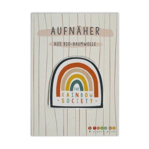 Può includere: Un toppa termoadesiva a forma di arcobaleno con il testo "The Rainbow Society" su sfondo bianco. La toppa è su una carta con il testo "Aufnäher aus Bio-Baumwolle" e "Tell Me organic goods".