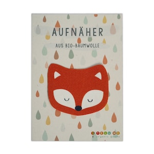 Op de afbeelding: Oranje vilt vos applique met gesloten ogen. De applique is op een kaart met de tekst "AUFNÄHER AUS BIO-BAUMWOLLE" en een kleurrijk regenboogpatroon. De kaart heeft ook de tekst "TELL ME organic goods".