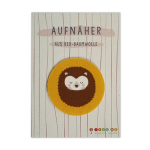 Könnte beinhalten: Ein gelber Stoff-Aufnäher mit einem braunen Igelgesicht darauf. Der Aufnäher trägt die Aufschrift "AUFNÄHER AUS BIO-BAUMWOLLE" und "TELL ME organic goods".