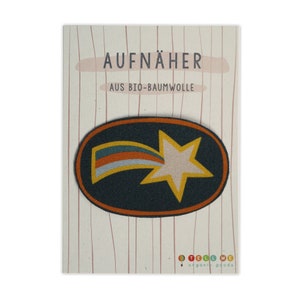 Könnte beinhalten: Ein dunkelgrüner ovaler Stoff-Aufnäher mit orangefarbenem Rand. Der Aufnäher zeigt einen gelben Stern mit weißem Zentrum und einen regenbogenfarbenen Sternschnuppen. Der Text "AUFNÄHER AUS BIO-BAUMWOLLE" ist auf der Karte gedruckt.