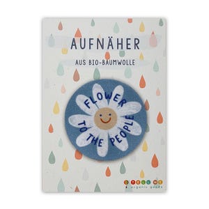 Op de afbeelding: Een blauwe en witte geborduurde strijk-op patch met een madeliefje en de tekst "Flower to the People". De patch is op een witte kaart met de tekst "Aufnäher aus Bio-Baumwolle" en een logo voor Tell Me Organic Goods.