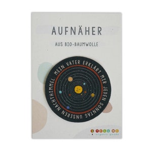 Peut inclure: Un patch rond en tissu noir avec une illustration colorée du système solaire. Le texte autour du bord dit "Mein Vater erklärt mir jeden Sonntag unseren Nachthimmel". Le patch est étiqueté "Aufnäher" et "Aus Bio-Baumwolle".
