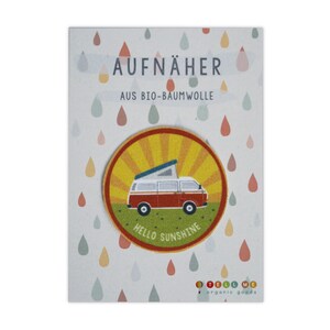 May include: A round, yellow, orange and green iron-on patch featuring a red and white camper van with a pop-up roof. The patch has the text "Hello Sunshine" in a yellow circle. The patch is on a white card with the text "Aufnäher aus Bio-Baumwolle" and "Tell Me organic goods".