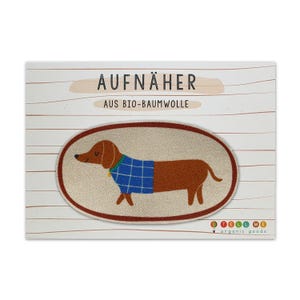 Pode incluir: Um remendo oval branco com um dachshund marrom vestindo um suéter azul e branco xadrez, com o texto "AUFNÄHER AUS BIO-BAUMWOLLE" e o logotipo "TELL ME organic goods".