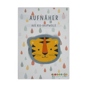 May include: A blue and yellow tiger face iron-on patch with the text "AUFNÄHER AUS BIO-BAUMWOLLE" on a white background with colorful raindrops.