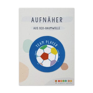 Peut inclure: Un patch en tissu bleu avec un ballon de football blanc et le texte "TEAM PLAYER" en cercle. Le patch est fait de coton biologique et est destiné à être cousu sur des vêtements.